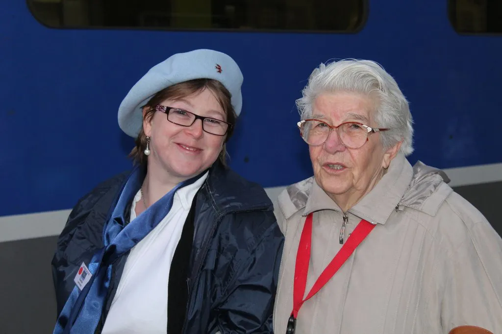 Une hospitalière en béret et une pèlerine âgée, joue contre joue, devant le train