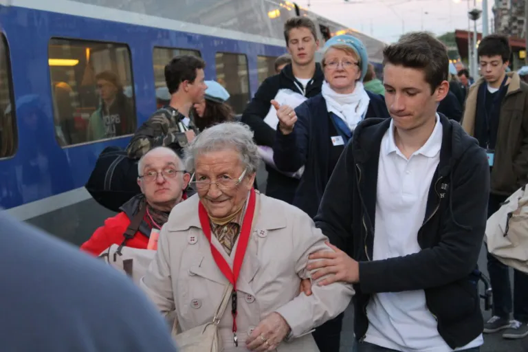Une pèlerine âgée entourée de jeunes hospitaliers souriants à bord du train